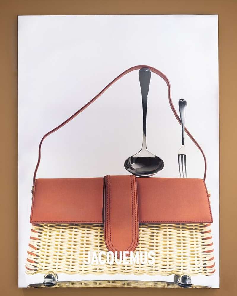 Promotional product shot featuring a brown and tan wicker handbag with a brown strap, accompanied by a large silver ladle and fork.
