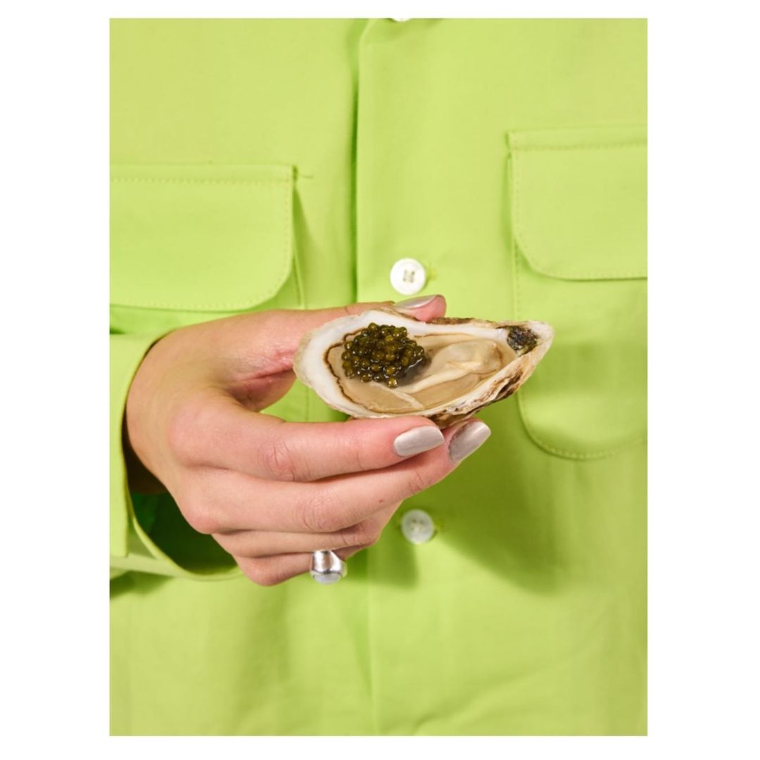 Close-up of a woman holding an oyster topped with caviar, showcasing food photography against a vibrant green shirt.
