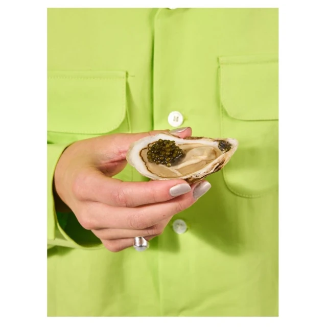Close-up of a woman holding an oyster topped with caviar, showcasing food photography against a vibrant green shirt.