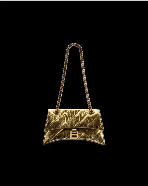 Gold Balenciaga purse with chain strap on a black backdrop. Metallic, shimmering material with a distinctive 'B' logo clasp.