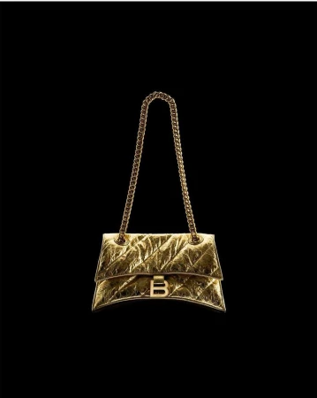 Gold Balenciaga purse with chain strap on a black backdrop. Metallic, shimmering material with a distinctive 'B' logo clasp.