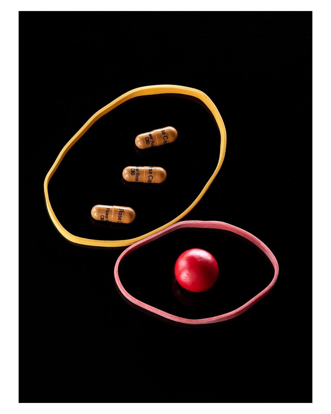 Pills inside yellow & pink rubber bands on a black surface. One red sphere sits inside the pink band. The capsules have text on them.