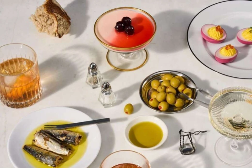 Overhead shot of a table arrangement featuring cocktails, deviled eggs, olives, and canned fish with warm lighting, inviting culinary presentation.