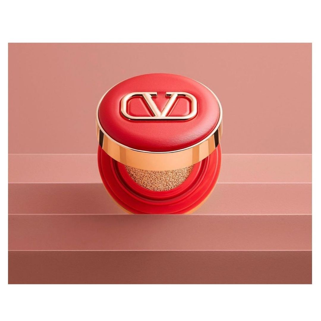 Cushion foundation in red packaging with a gold logo detail sits on a multi-tiered pink surface in a studio setting. Cosmetic product.