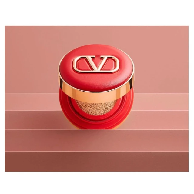 Cushion foundation in red packaging with a gold logo detail sits on a multi-tiered pink surface in a studio setting. Cosmetic product.