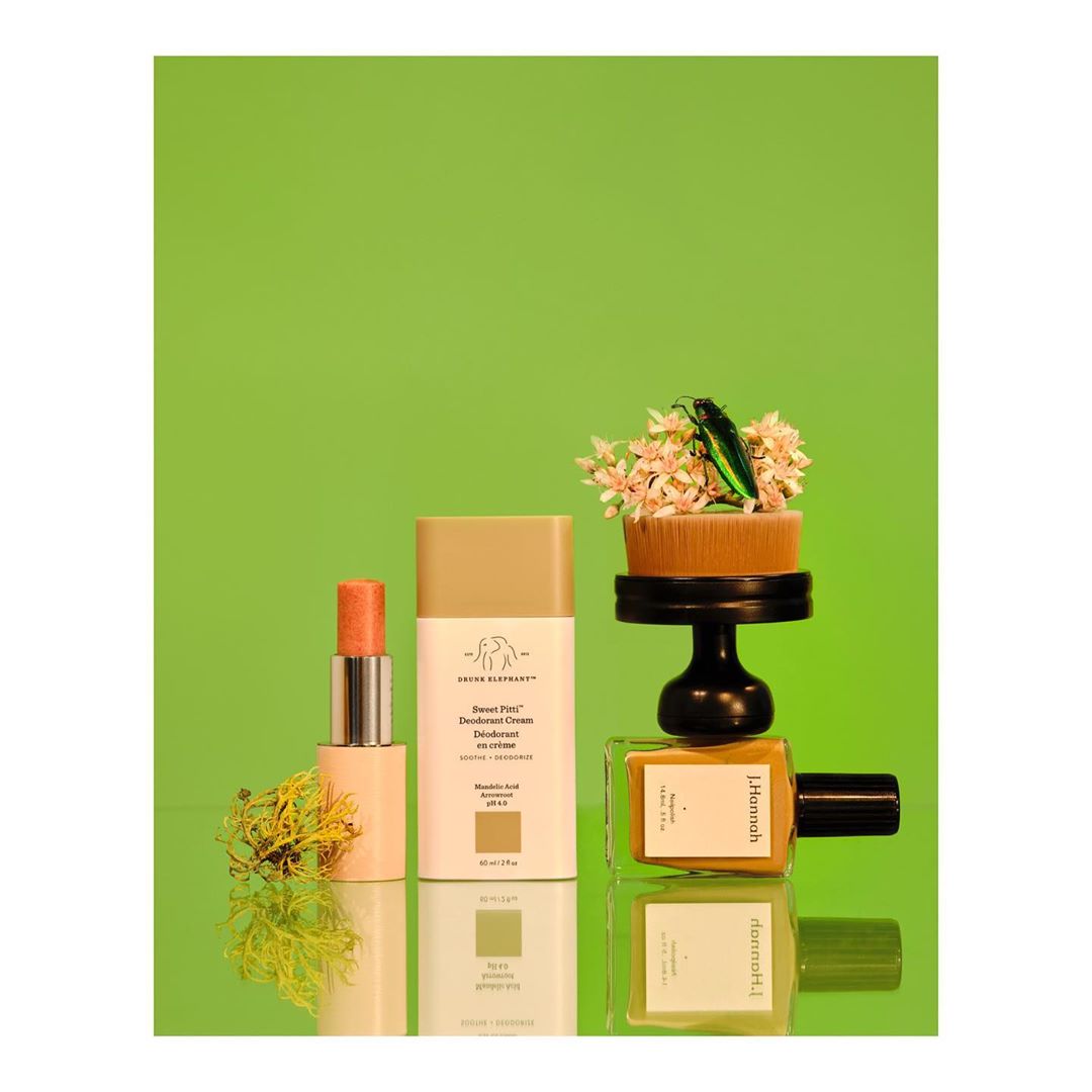 Cosmetic product still life featuring lip balm, deodorant, nail polish, and floral arrangement against a vibrant green backdrop.