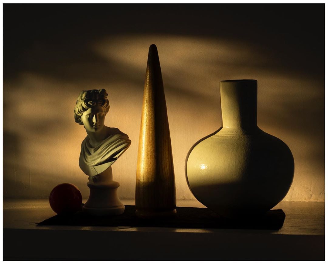 Still life shot of a bust statue, cone, vase, and ball arranged together, lit with warm, directional light in a dark indoor setting.