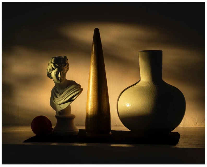 Still life shot of a bust statue, cone, vase, and ball arranged together, lit with warm, directional light in a dark indoor setting.