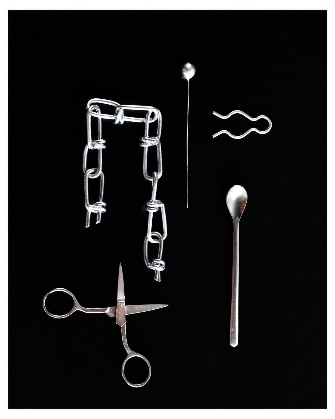 Still life of a silver chain, hairpin, stickpin, spoon, and scissors against a black background, showcasing metal objects.