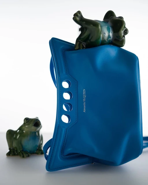 Blue designer handbag with ceramic frog figurines, placed against a bright white backdrop in a fashion studio setting.