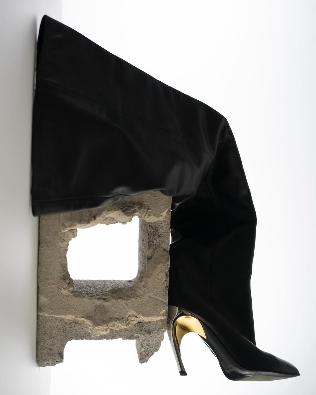 Black leather thigh-high boot with a gold metallic heel is balanced on a weathered cinder block against a clean white background.
