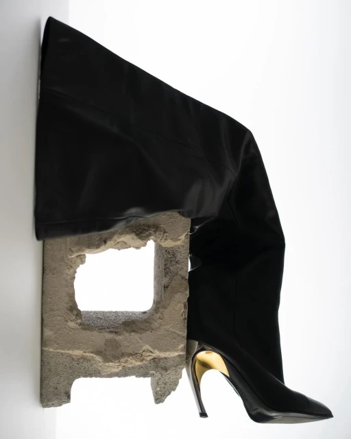 Black leather thigh-high boot with a gold metallic heel is balanced on a weathered cinder block against a clean white background.