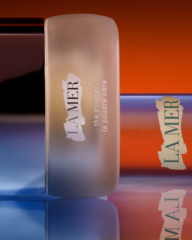 La Mer 'the powder' loose powder container in front of an abstract orange and blue studio background with a reflective surface.