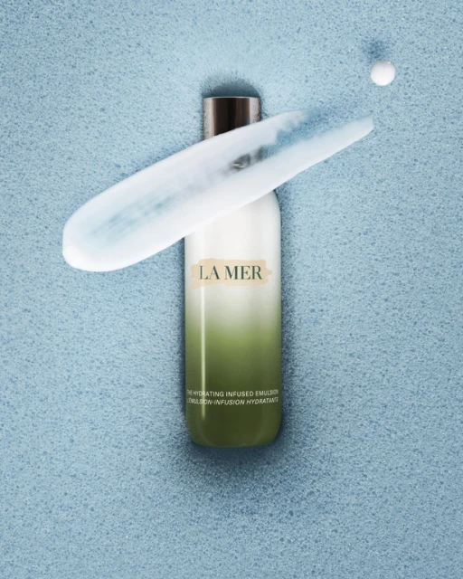 Elevated view of La Mer hydrating emulsion bottle with dab of lotion on a light blue textured surface, perfect for skincare ad campaign.