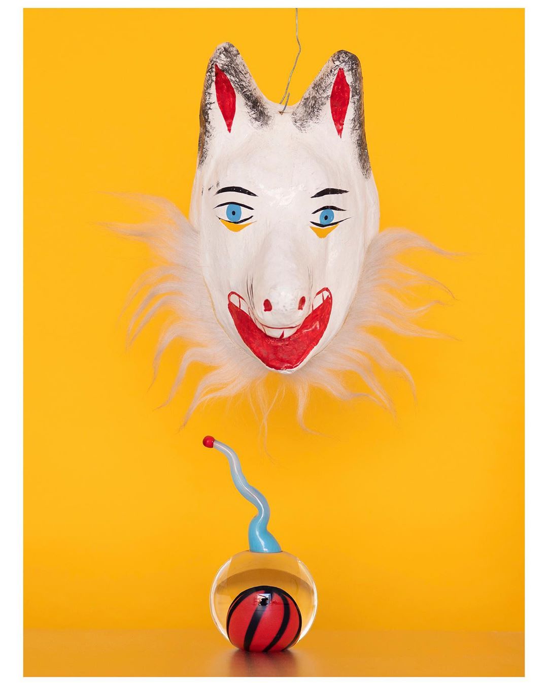 Close-up still life of a hanging paper mache mask with white fur collar, above a transparent sphere holding a striped ball.
