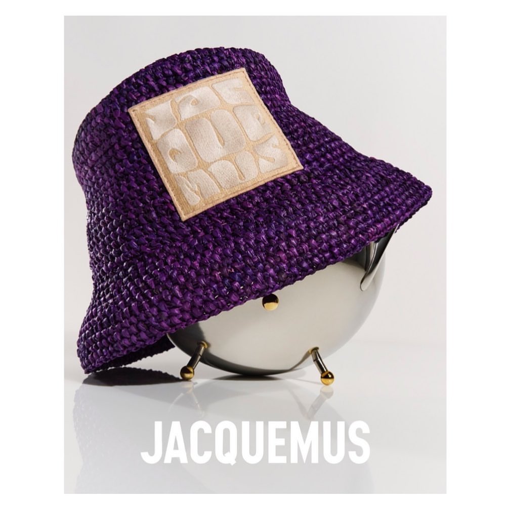 Purple straw bucket hat with a tan square patch sits atop a polished silver bowl. The brand name JACQUEMUS is centered below.