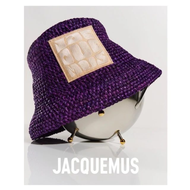 Purple straw bucket hat with a tan square patch sits atop a polished silver bowl. The brand name JACQUEMUS is centered below.