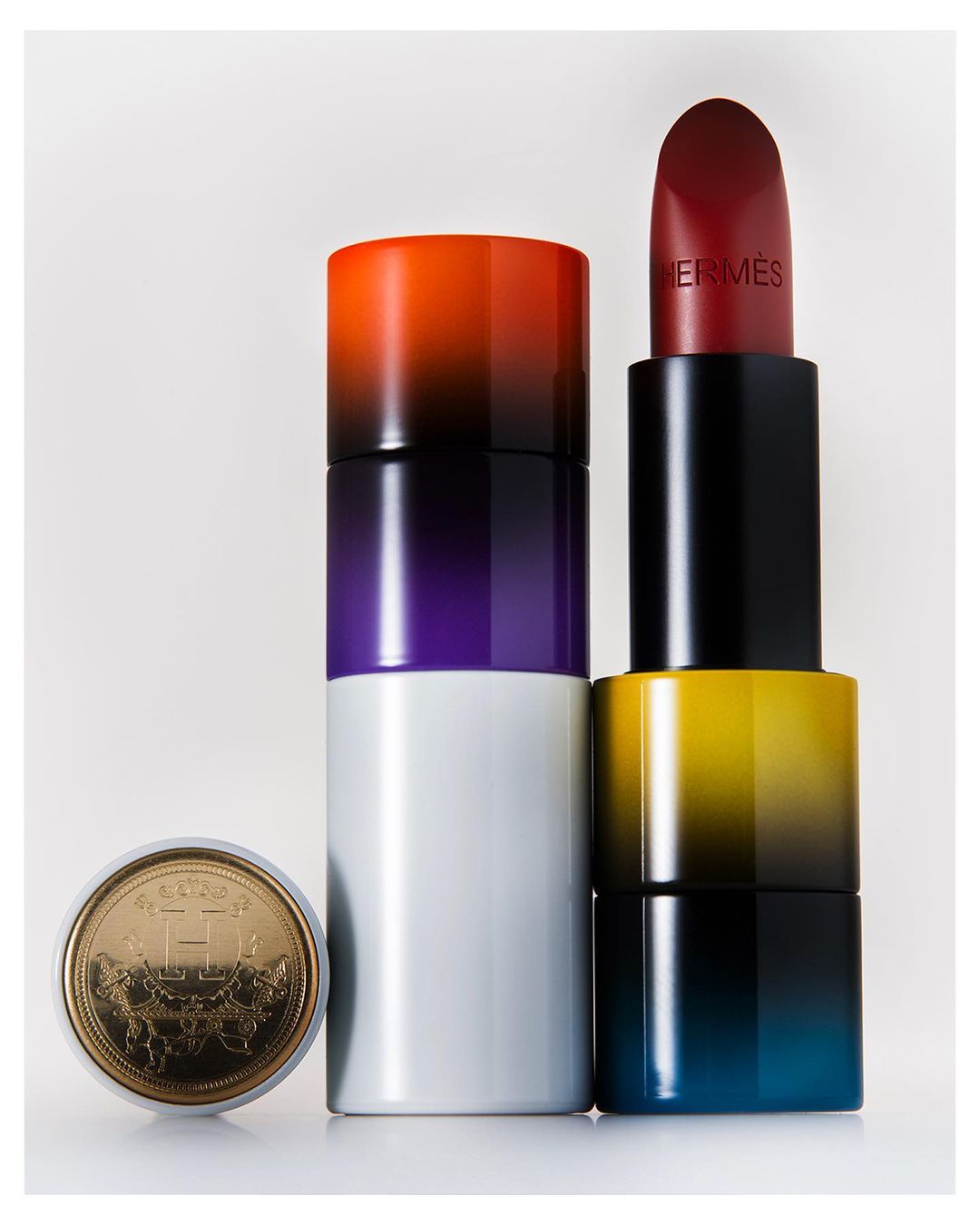 Elevated product shot of Hermès lipstick and gold coin with Hermès logo, showcasing luxury beauty and brand recognition on white.