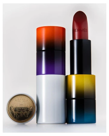 Elevated product shot of Hermès lipstick and gold coin with Hermès logo, showcasing luxury beauty and brand recognition on white.