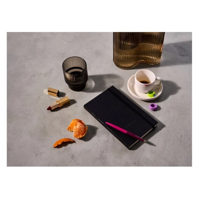 Still life photo of a black notebook, pink pen, lipstick, glass vases, peeled tangerine, and coffee cup on a textured gray surface.