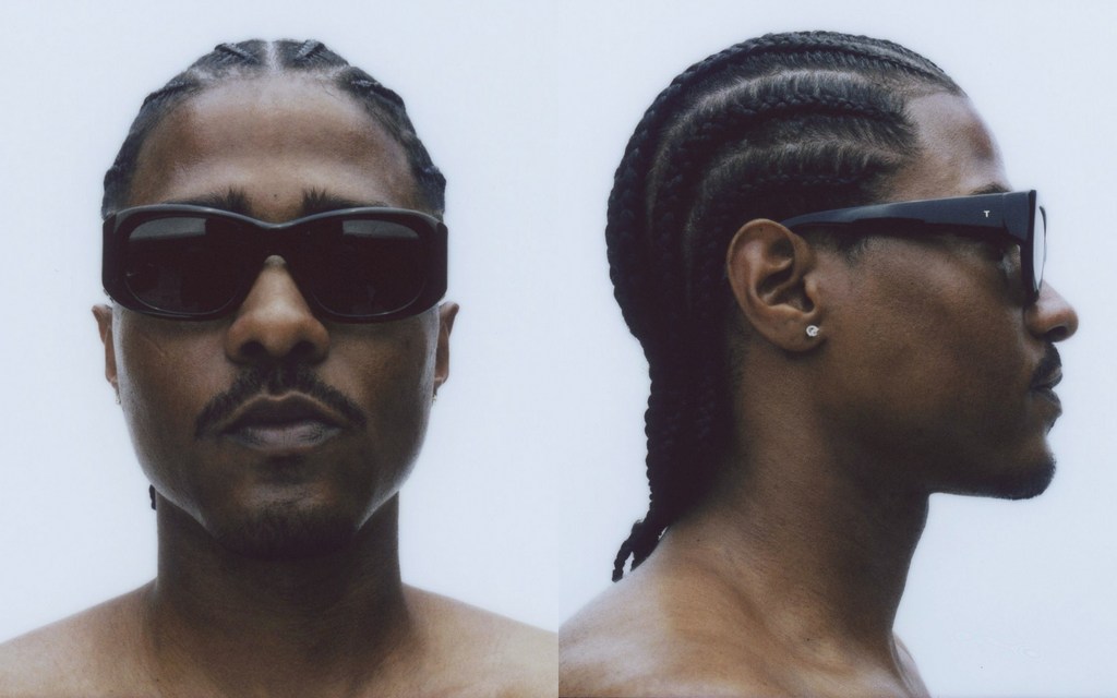 Portrait of an adult man with braided hair and a moustache, wearing sunglasses and shot in front of a white backdrop.