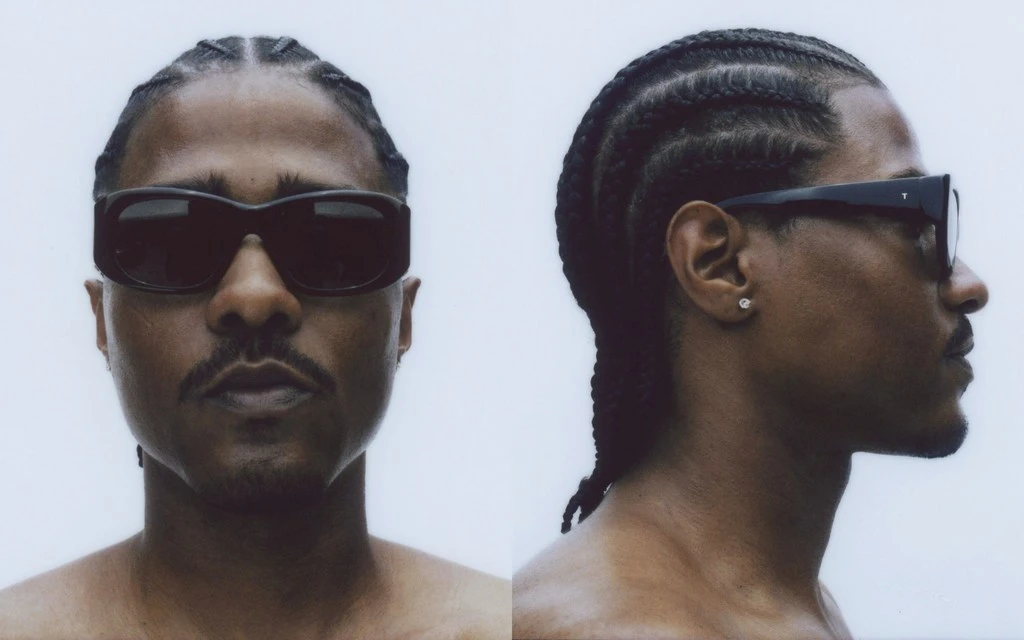 Portrait of an adult man with braided hair and a moustache, wearing sunglasses and shot in front of a white backdrop.