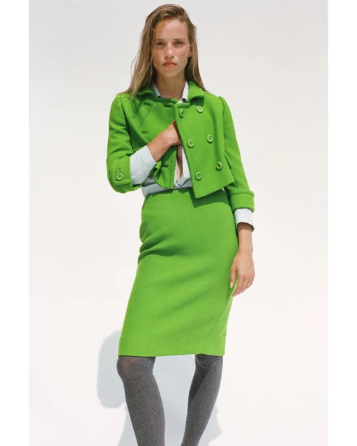 Fashion editorial shot of a woman wearing a green skirt suit, posing in front of a white background, styled with grey tights and light makeup.