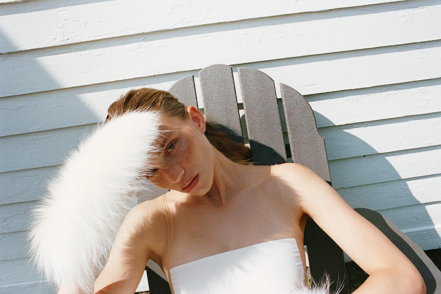 A woman is sitting in a chair in front of a white house, wearing a white top and a white furry stole. The sun is shining on her face.