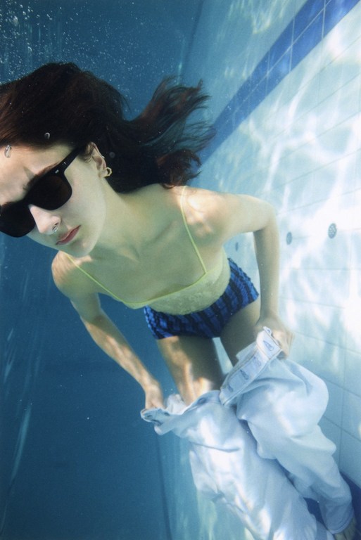Brunette woman underwater in a swimming pool wearing sunglasses, shorts, and a shirt, creates an ethereal and serene image.