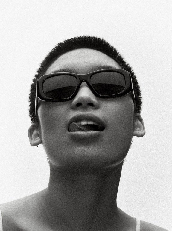Close-up monochrome portrait of a person with short hair wearing black sunglasses and sticking out their tongue against a white background