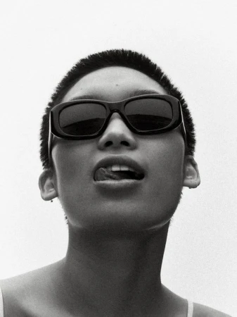 Close-up monochrome portrait of a person with short hair wearing black sunglasses and sticking out their tongue against a white background