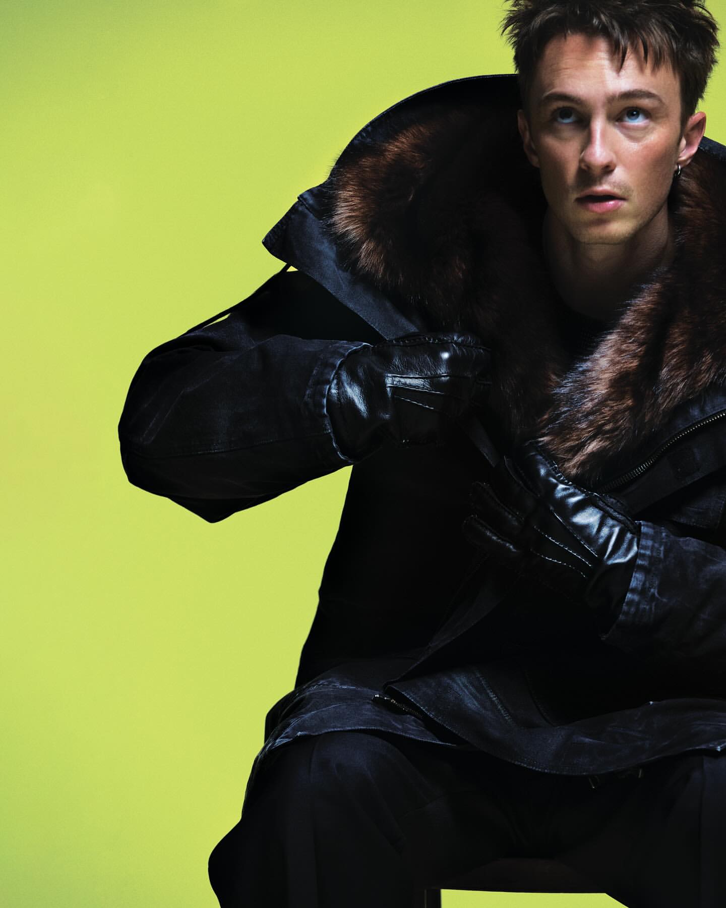Man in a fur-collared coat and leather gloves sits against a bright yellow background, looking up and to the right in a fashion shot.