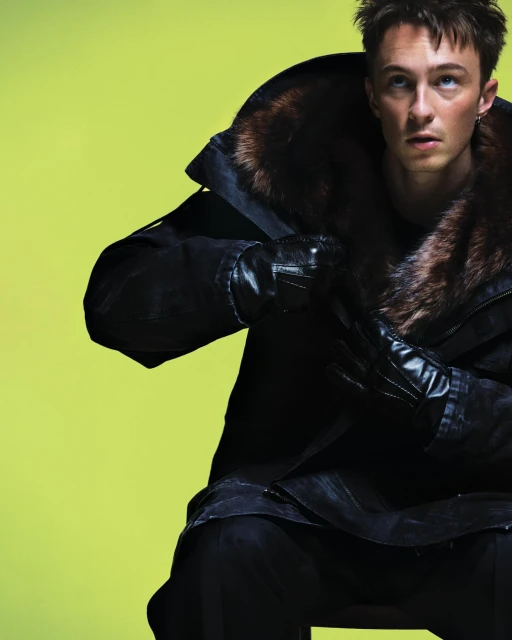 Man in a fur-collared coat and leather gloves sits against a bright yellow background, looking up and to the right in a fashion shot.