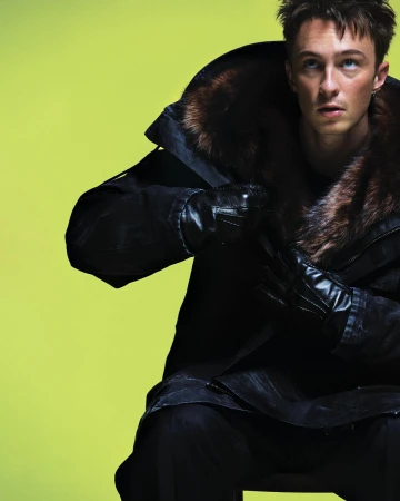 Man in a fur-collared coat and leather gloves sits against a bright yellow background, looking up and to the right in a fashion shot.