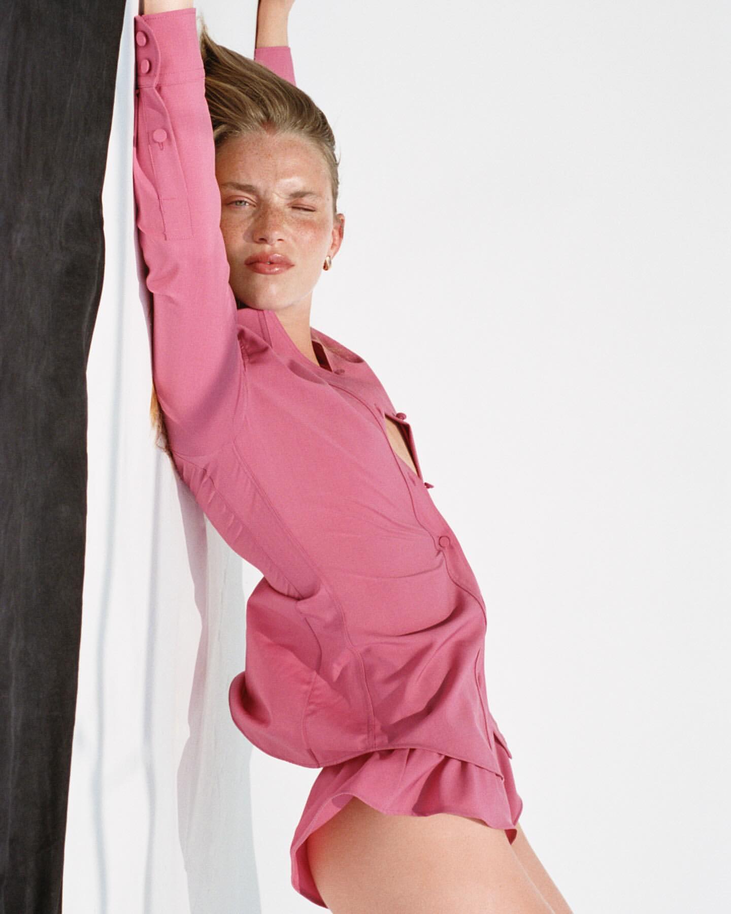 Woman with fair skin leans against a two-tone wall in a pink button-down top and shorts in a studio with soft lighting.