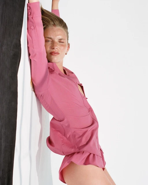 Woman with fair skin leans against a two-tone wall in a pink button-down top and shorts in a studio with soft lighting.