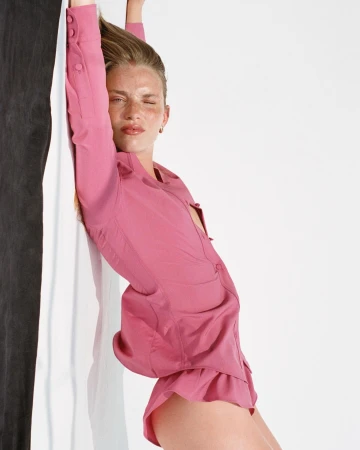 Woman with fair skin leans against a two-tone wall in a pink button-down top and shorts in a studio with soft lighting.