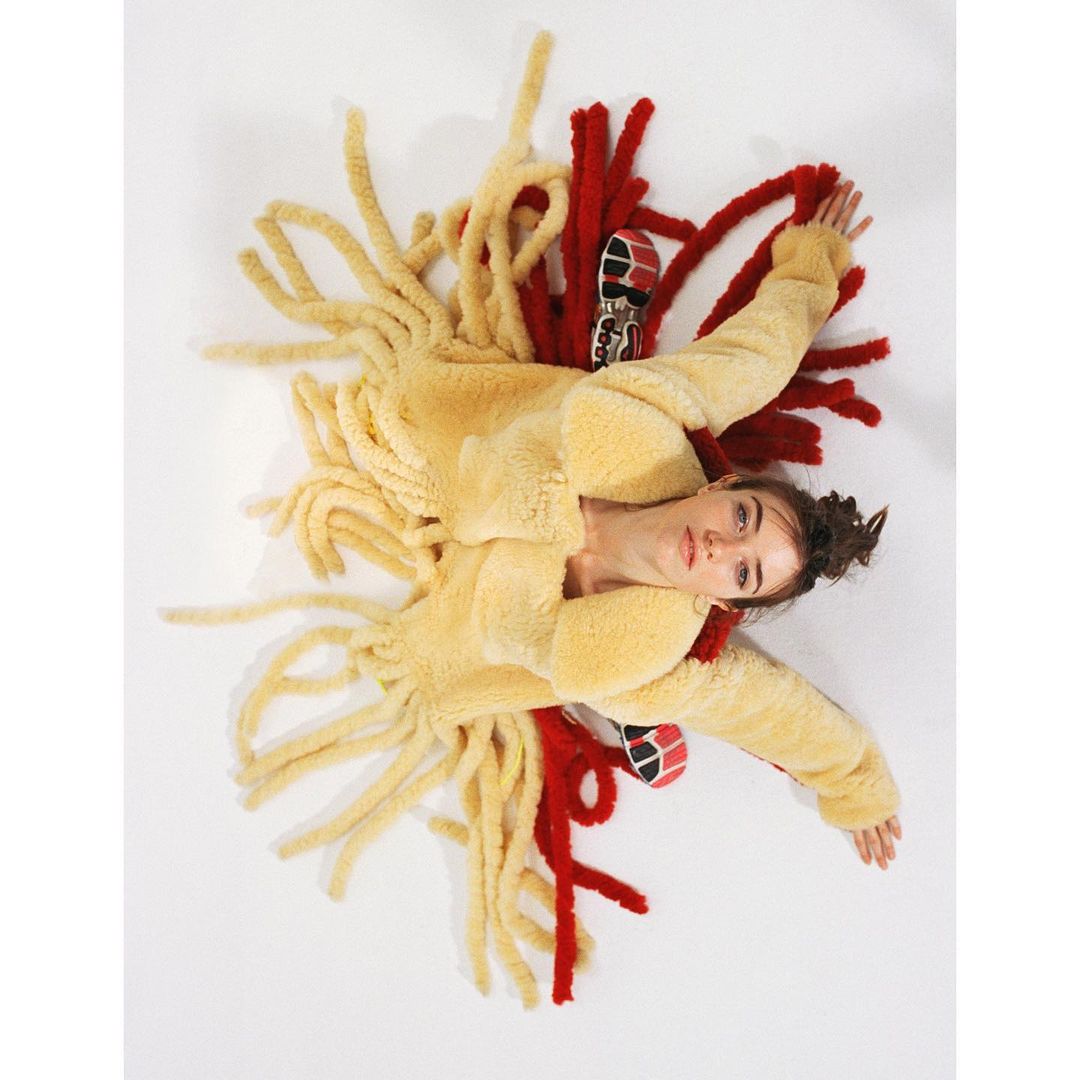 Overhead shot of a young woman lying on a white surface wearing a beige fringe jacket with red fringe details and silver sneakers.
