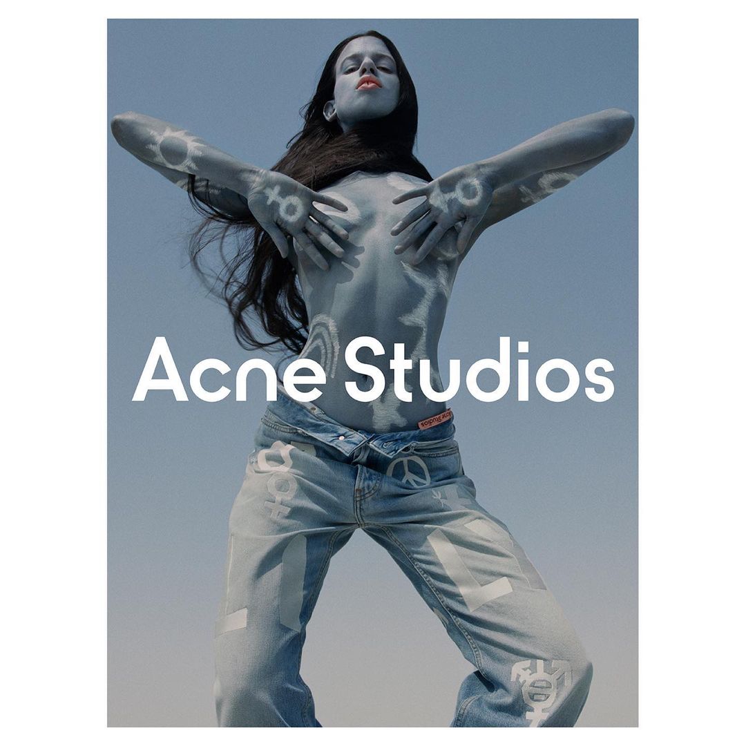 Woman poses with body paint and jeans in a studio shot, showcasing a fashion-forward look with visible branding and peace symbols.