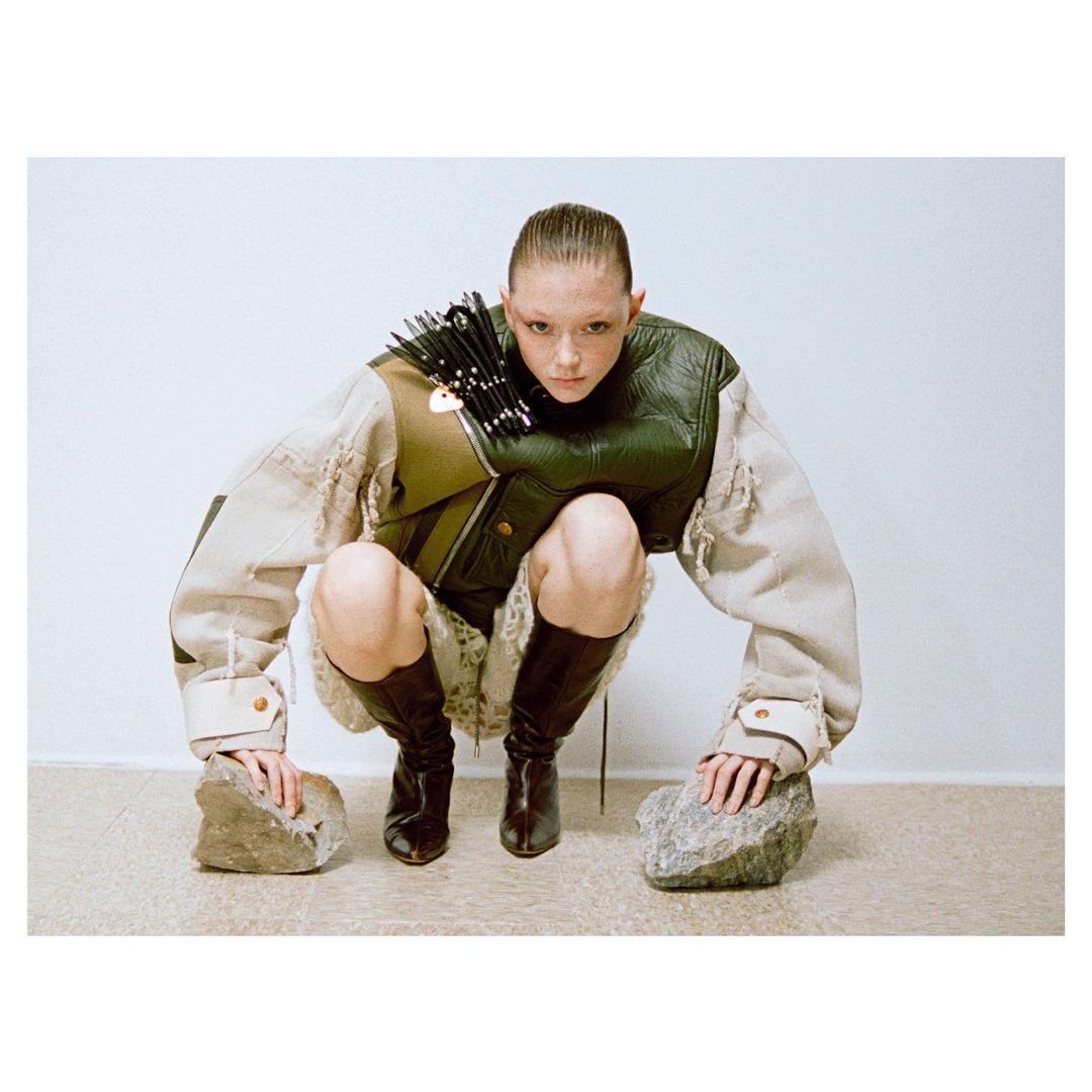 Model squatting in a structured green and cream jacket and dark boots, touching rocks on the floor against a plain wall.