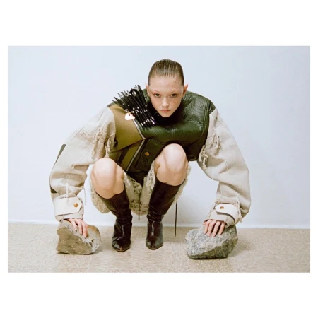 Model squatting in a structured green and cream jacket and dark boots, touching rocks on the floor against a plain wall.