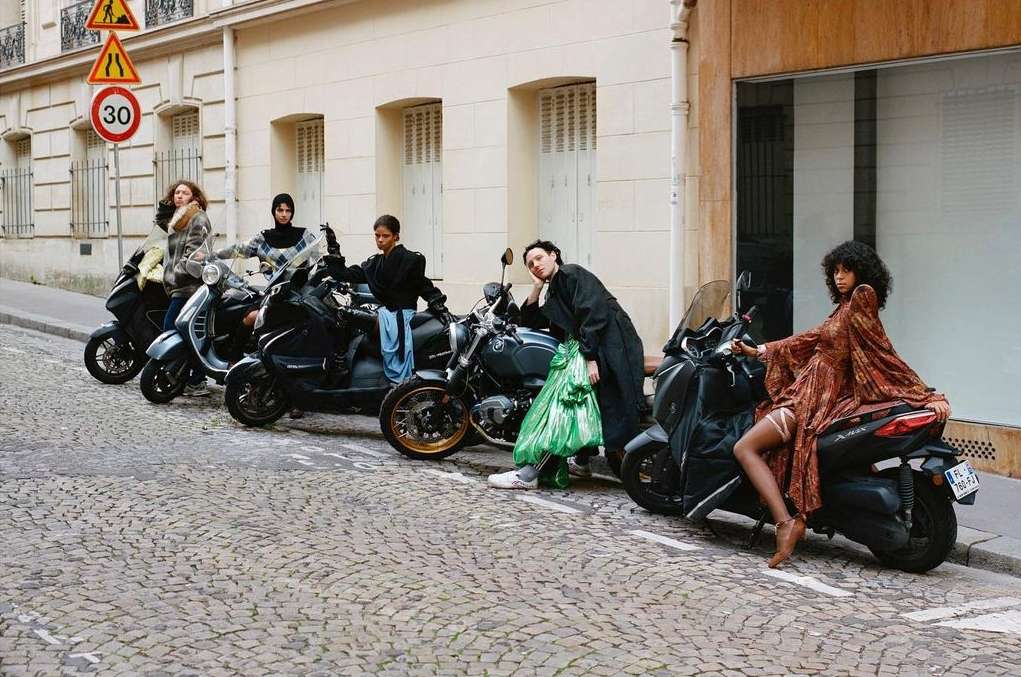 Group of stylish adult women pose on scooters and motorcycles on a city street, wearing fashionable clothing and accessories.
