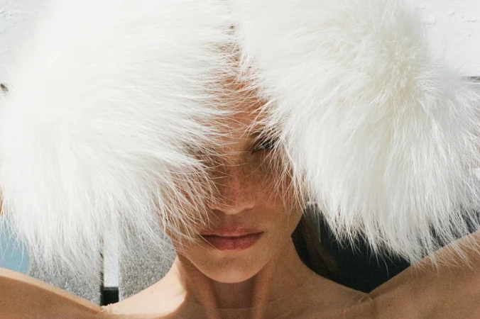 Close-up portrait of a woman wearing a large white fluffy faux fur hat framing her face, photographed by Thistle Brown.