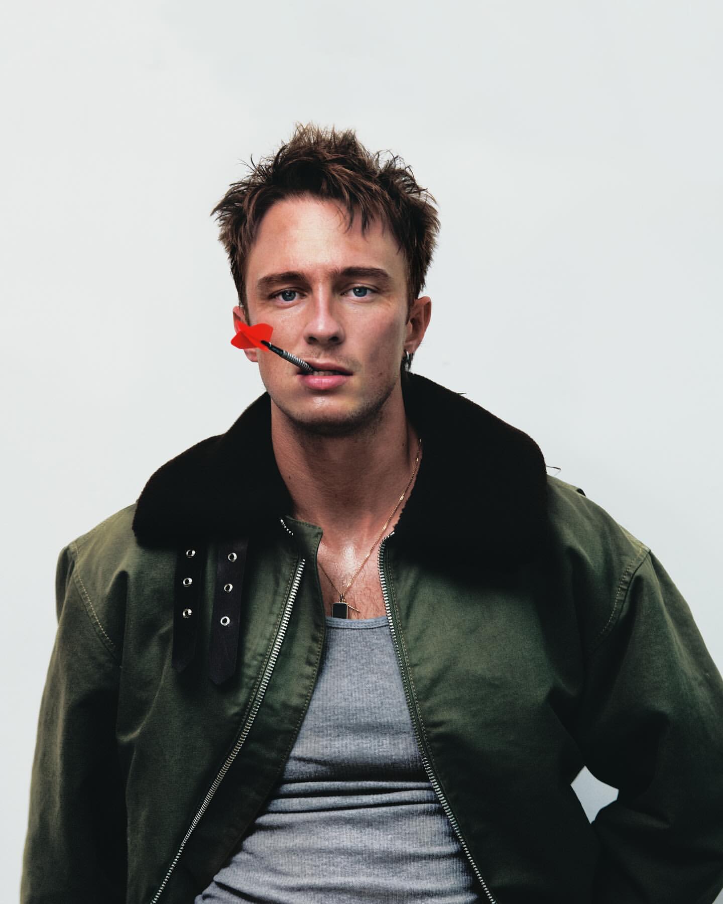 Stylish portrait of a young man wearing a green military jacket with a red dart in his mouth against a white background in studio.