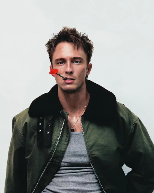 Stylish portrait of a young man wearing a green military jacket with a red dart in his mouth against a white background in studio.