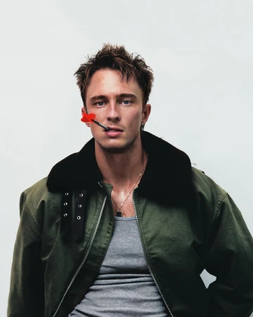 Stylish portrait of a young man wearing a green military jacket with a red dart in his mouth against a white background in studio.