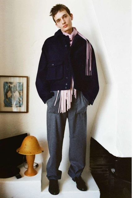 Young man in a navy jacket, pink shirt, and gray pants poses indoors against a white wall with an orange lamp and framed artwork.