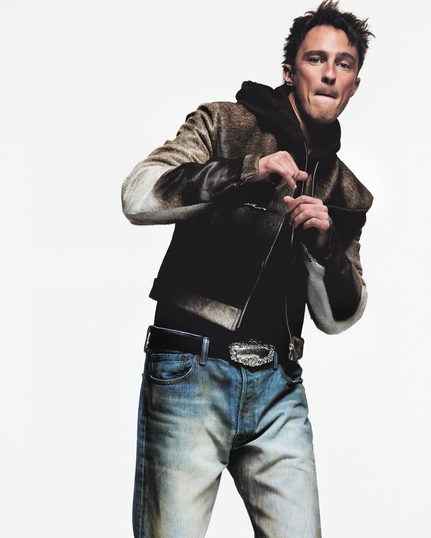 A fashionable adult male poses indoors, wearing a hooded jacket, jeans, and decorative belt. The image features a clean white backdrop.