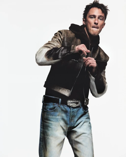 A fashionable adult male poses indoors, wearing a hooded jacket, jeans, and decorative belt. The image features a clean white backdrop.