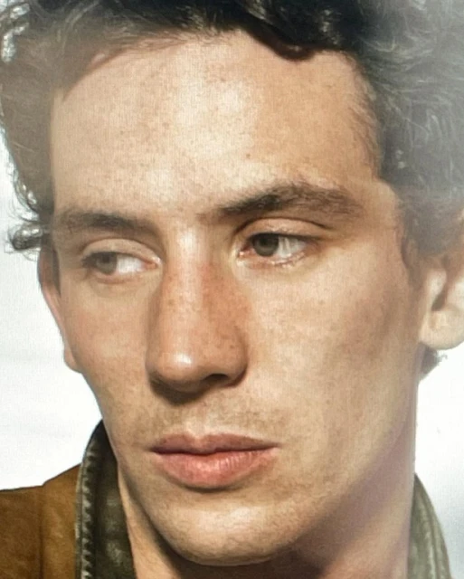 Close-up studio shot of a young adult male with freckles, dark curly hair, wearing a tan jacket and looking left.